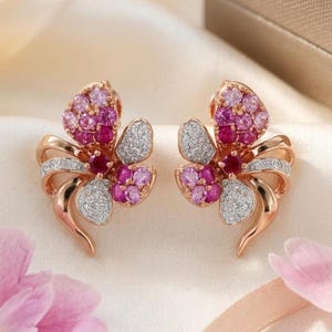 May include: A pair of rose gold earrings in a flower design. Each earring has pink and purple gemstones, a central ruby, and white gemstone accents. The earrings have a curved, ribbon-like shape, with a delicate appearance.