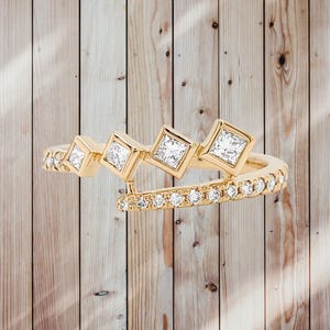 Puede incluir: Un anillo de oro con tres diamantes de talla cuadrada y una banda de diamantes más pequeños. El anillo tiene un diseño abierto único, con la banda que se envuelve alrededor del dedo. El anillo está sobre un fondo de madera.