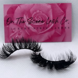 May include: A pair of black false eyelashes, with a long, wispy, and dramatic look. The eyelashes are on a white background, with a pink card behind them that says "On The Scene Lash Co. Luxury Strip Lashes".