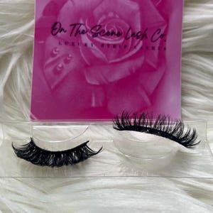 May include: A pair of black false eyelashes in a clear plastic container. The lashes are long and full, with a natural-looking curl. A pink card with the text "On The Scene Lash Co. LUXURY STRIP LASHES" is in the background.