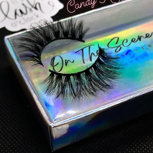 K&ouml;nnte beinhalten: Ein Paar schwarze falsche Wimpern in einer holografischen Schachtel. Auf der Schachtel steht "On The Scene" und "Candy Kisses 18mm". Ein Aufkleber mit dem Text "It's Lash O'Clock" ist ebenfalls sichtbar.