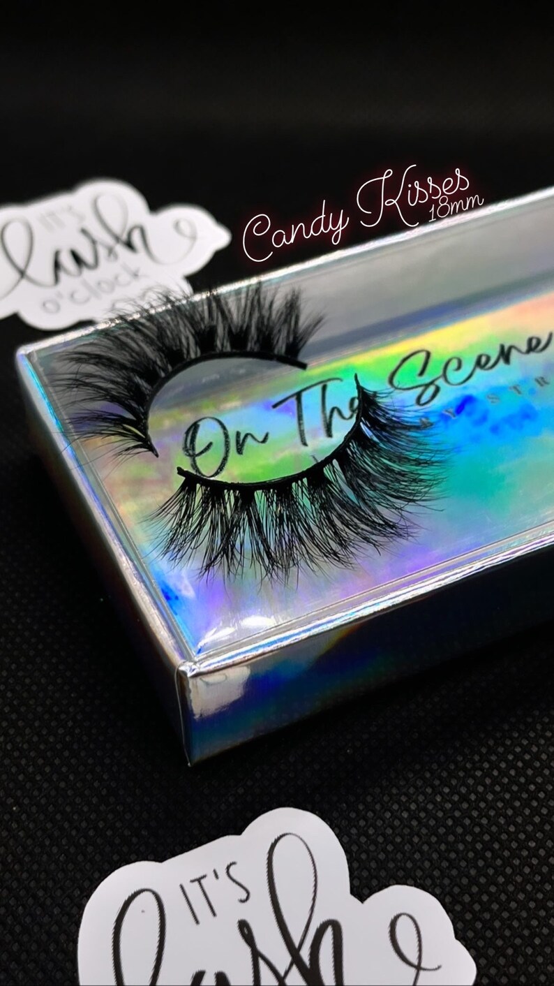 K&ouml;nnte beinhalten: Ein Paar schwarze falsche Wimpern in einer holografischen Schachtel. Auf der Schachtel steht "On The Scene" und "Candy Kisses 18mm". Ein Aufkleber mit dem Text "It's Lash O'Clock" ist ebenfalls sichtbar.