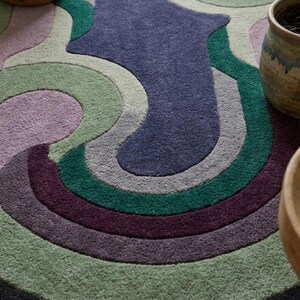 Puede incluir: Una alfombra redonda y multicolor con un dise&ntilde;o retro. La alfombra presenta una serie de formas curvas y superpuestas en tonos de morado, verde, gris y azul. La alfombra est&aacute; bordeada por un borde gris oscuro. Dos macetas de cer&aacute;mica son visibles en el fondo.
