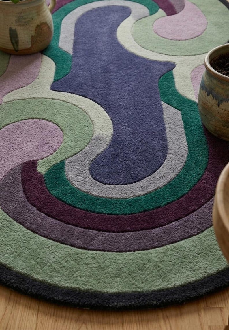 Puede incluir: Una alfombra redonda y multicolor con un dise&ntilde;o retro. La alfombra presenta una serie de formas curvas y superpuestas en tonos de morado, verde, gris y azul. La alfombra est&aacute; bordeada por un borde gris oscuro. Dos macetas de cer&aacute;mica son visibles en el fondo.