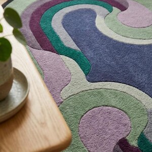 Puede incluir: Una alfombra redonda con un dise&ntilde;o ondulado en tonos azules, verdes, morados y grises. La alfombra tiene un borde negro y est&aacute; hecha de un material suave y texturizado. La alfombra est&aacute; colocada sobre un suelo de madera.
