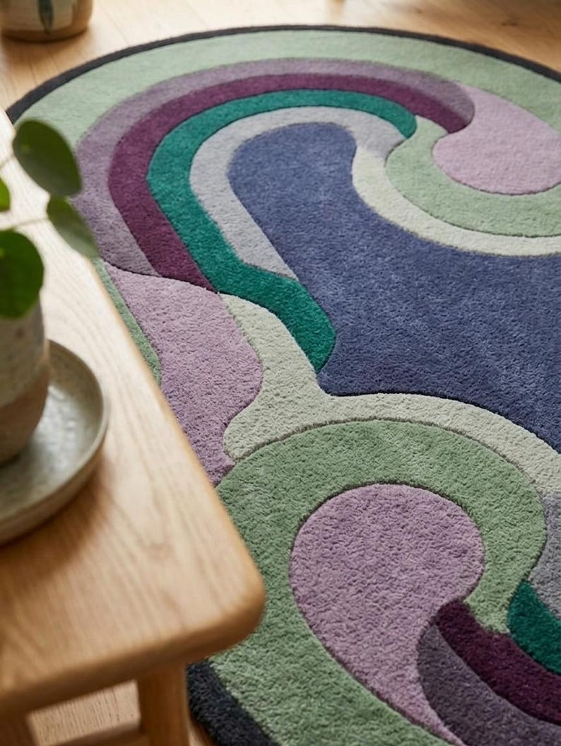 Puede incluir: Una alfombra redonda con un dise&ntilde;o ondulado en tonos azules, verdes, morados y grises. La alfombra tiene un borde negro y est&aacute; hecha de un material suave y texturizado. La alfombra est&aacute; colocada sobre un suelo de madera.