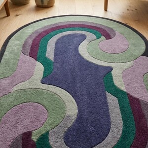 Puede incluir: Alfombra ovalada con un dise&ntilde;o abstracto y colorido. La alfombra presenta una forma central azul oscuro rodeada de patrones arremolinados en tonos de morado, verde y gris. La alfombra est&aacute; sobre un suelo de madera.