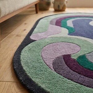 Puede incluir: Una alfombra ovalada con dise&ntilde;o retro. Presenta bandas curvas en tonos verdes, morados y azules, con un borde gris oscuro. La alfombra est&aacute; hecha de un material suave y texturizado, colocada sobre un suelo de madera.