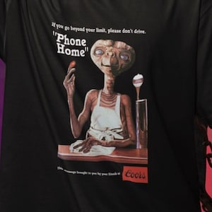 May include: Black t-shirt with a graphic print of an alien character from the movie E.T. The Extra-Terrestrial. The alien is holding a piece of candy and the text "Phone Home" is printed above the alien. The text "If you go beyond your limit, please don't drive." is printed above the alien. The text "Coors" is printed below the alien.