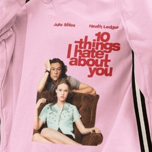 Peut inclure: Sweat-shirt rose avec un imprimé graphique du film "10 choses que je déteste chez toi" mettant en scène Julia Stiles et Heath Ledger. Le texte sur le sweat-shirt est en rouge.