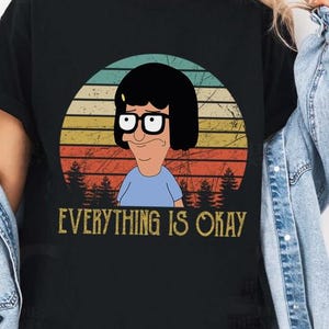 Op de afbeelding: Een zwart T-shirt met korte mouwen en een afbeelding van een stripfiguur met zwart haar en een bril, gecentreerd in een cirkelvormige zonsondergangachtergrond met silhouetten van dennenbomen. De tekst "EVERYTHING IS OKAY" staat in het geel eronder.