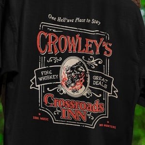 Pode incluir: Camiseta preta com um design gr&aacute;fico em estilo vintage. O design apresenta o texto "Crowley's Crossroads Inn" em vermelho e branco, com texto adicional que diz "Fine Whiskey" e "Great Deals". Uma imagem circular de um dem&ocirc;nio est&aacute; no centro.
