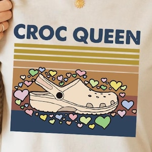 May include: A beige t-shirt with the text "CROC QUEEN" in blue letters. The shirt features a vintage-style design with a brown and yellow stripe and a white and beige cartoon image of a clog shoe surrounded by colorful hearts. The text "Sand" is printed in black at the bottom of the shirt.