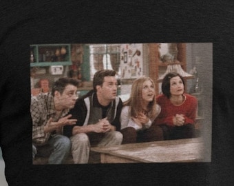 May include: Black t-shirt featuring a rectangular image of the cast of the TV show Friends. The image shows four people sitting and looking upwards. The t-shirt is made of a soft material.