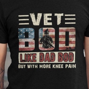 Op de afbeelding: Zwart t-shirt met de tekst "VET BOD" in witte letters boven een afbeelding van de Amerikaanse vlag met een silhouet van een soldaat. Daaronder staat de tekst "LIKE DAD BOD BUT WITH MORE KNEE PAIN" in rode letters.