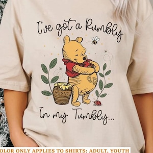 May include: A sand-colored short-sleeve t-shirt featuring a graphic of Winnie the Pooh holding a honey pot. The text reads, "I've got a Rumbly in my Tumbly..." with small illustrations of a bee, a ladybug, and green leafy stems surrounding the character.