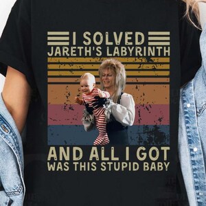 May include: A black short-sleeved t-shirt featuring a graphic print of a person holding a baby. The text reads I SOLVED JARETH'S LABYRINTH AND ALL I GOT WAS THIS STUPID BABY, set against a retro-style background of horizontal stripes in muted colors.