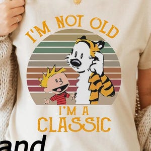 May include: Sand-colored t-shirt featuring a graphic of Calvin and Hobbes with the text "I'M NOT OLD I'M A CLASSIC". The design includes a circular graphic with a striped background. The shirt is a short-sleeved crew neck.