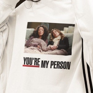 May include: White sweatshirt with a graphic print of two women in a hospital bed. The text "You're My Person" is printed below the image.