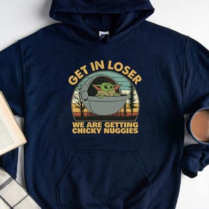 May include: Navy blue hoodie featuring a graphic of Grogu in a pod with the text "GET IN LOSER WE ARE GETTING CHICKY NUGIES". The design is in a retro style with a sunset background. A mug of coffee and a book are also in the image.