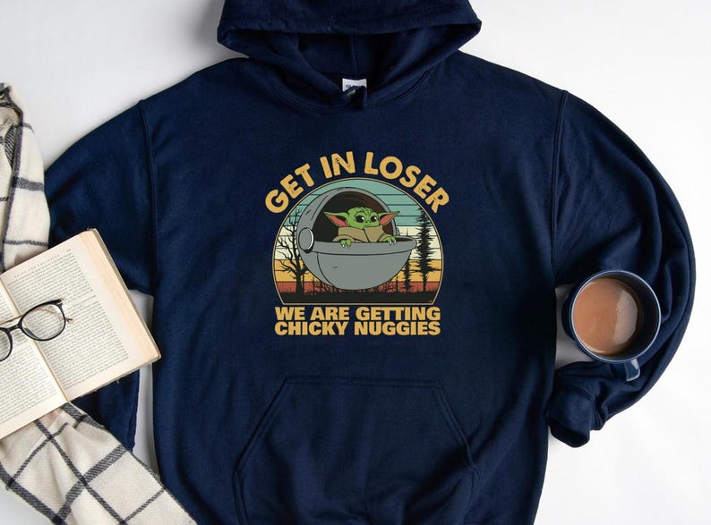 May include: Navy blue hoodie featuring a graphic of Grogu in a pod with the text "GET IN LOSER WE ARE GETTING CHICKY NUGIES". The design is in a retro style with a sunset background. A mug of coffee and a book are also in the image.