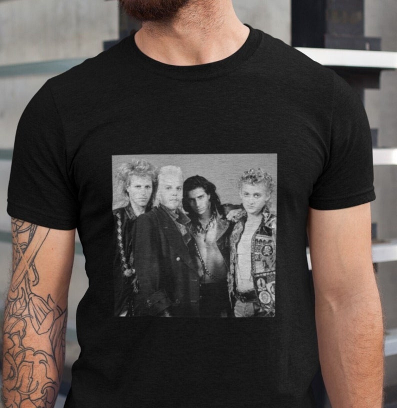 Op de afbeelding: Zwart-witfoto van vier mannen in een band. De mannen dragen leren jassen en jeans. De foto is op een zwart T-shirt.