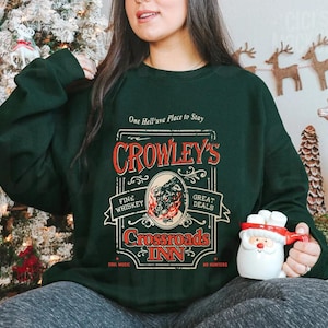 Pode incluir: Camisola verde escuro com o texto "One Hell'uva Place to Stay" e "Crowley's Crossroads Inn" em vermelho e dourado. O design inclui um lobo e chamas. A pessoa est&aacute; segurando uma caneca do Papai Noel.
