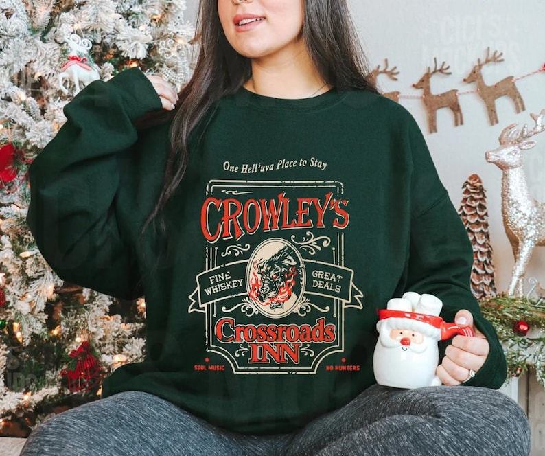 Pode incluir: Camisola verde escuro com o texto "One Hell'uva Place to Stay" e "Crowley's Crossroads Inn" em vermelho e dourado. O design inclui um lobo e chamas. A pessoa est&aacute; segurando uma caneca do Papai Noel.