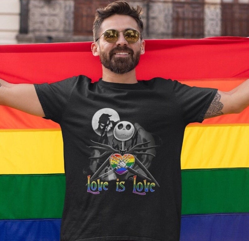May include: A black t-shirt with a graphic of Jack Skellington from The Nightmare Before Christmas holding a rainbow heart. The text "Love is Love" is printed below the graphic. The person wearing the shirt is standing in front of a rainbow pride flag.