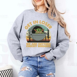 May include: A light gray sweatshirt with a graphic featuring a green alien character in a pod, with the text "GET IN LOSER" and "WE ARE GETTING CHICKY NUGGS". The design has a retro sunset and forest background.