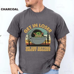 May include: Charcoal gray t-shirt featuring a graphic of Grogu in a pod with the text "GET IN LOSER WE ARE GETTING CHICKY NUGGIES" in a circular design. The design includes a sunset and trees.