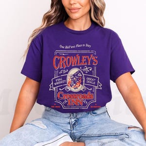 Pode incluir: Uma t-shirt roxa com um design gr&aacute;fico em estilo vintage para "Crowley's Crossroads Inn". O design inclui texto e uma ilustra&ccedil;&atilde;o de uma criatura semelhante a um lobo. A t-shirt tamb&eacute;m tem as frases "Fine Whiskey" e "Great Deals".