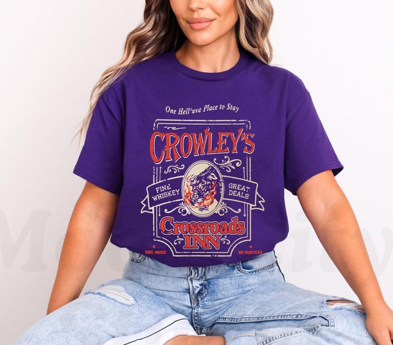 Pode incluir: Uma t-shirt roxa com um design gr&aacute;fico em estilo vintage para "Crowley's Crossroads Inn". O design inclui texto e uma ilustra&ccedil;&atilde;o de uma criatura semelhante a um lobo. A t-shirt tamb&eacute;m tem as frases "Fine Whiskey" e "Great Deals".
