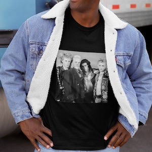 Op de afbeelding: Zwart T-shirt met een zwart-wit foto van een band met vijf leden. De bandleden dragen leren jacks en hebben lang haar. De foto is tegen een witte achtergrond.
