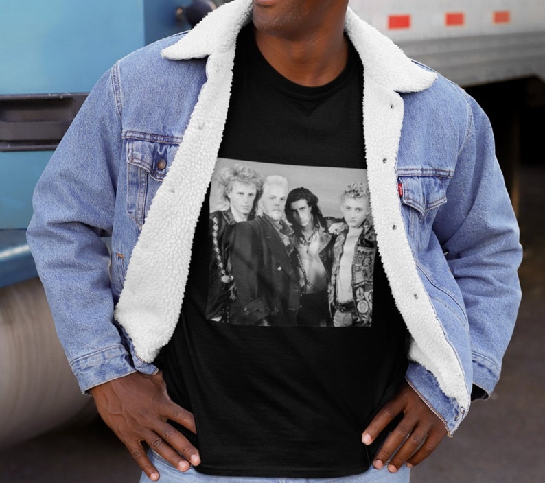 Op de afbeelding: Zwart T-shirt met een zwart-wit foto van een band met vijf leden. De bandleden dragen leren jacks en hebben lang haar. De foto is tegen een witte achtergrond.