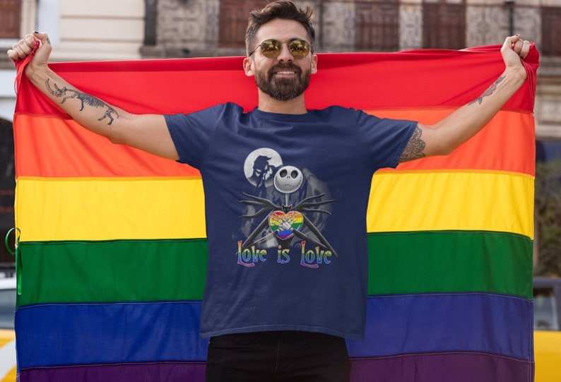 May include: A person wearing a navy blue t-shirt with a rainbow heart and the text "Love is Love" and a black and white illustration of Jack Skellington from The Nightmare Before Christmas. The person is holding a rainbow pride flag.