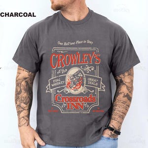 Pode incluir: Camiseta cinza carv&atilde;o com um gr&aacute;fico de estilo vintage. O gr&aacute;fico inclui o texto "CROWLEY'S Crossroads INN" em vermelho e branco, com uma ilustra&ccedil;&atilde;o de um lobo. A camisa tamb&eacute;m tem o texto "One Hell'uva Place to Stay."