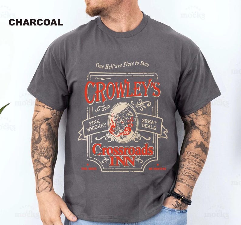 Pode incluir: Camiseta cinza carv&atilde;o com um gr&aacute;fico de estilo vintage. O gr&aacute;fico inclui o texto "CROWLEY'S Crossroads INN" em vermelho e branco, com uma ilustra&ccedil;&atilde;o de um lobo. A camisa tamb&eacute;m tem o texto "One Hell'uva Place to Stay."