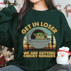 May include: Dark green sweatshirt with the text "GET IN LOSER" above a graphic of Baby Yoda in a pod. Below the graphic, the text reads "WE ARE GETTING CHICKY NUGGIES". The sweatshirt is a long-sleeved crew neck.