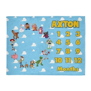 Op de afbeelding: Een lichtblauwe rechthoekige deken met een herhalend patroon van witte wolken. De deken toont de naam Axton in gele tekst, gevolgd door de cijfers 1 tot en met 12 en het woord Months. Diverse geanimeerde figuren zijn in een cirkel geplaatst.
