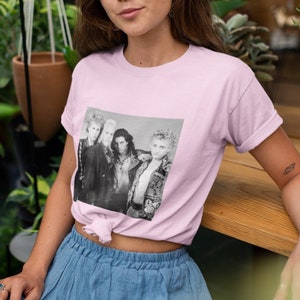 Op de afbeelding: Een roze T-shirt met een zwart-witfoto van een band op de voorkant. De bandleden dragen leren jacks en hebben lang haar. De foto is omlijst met een witte rand.