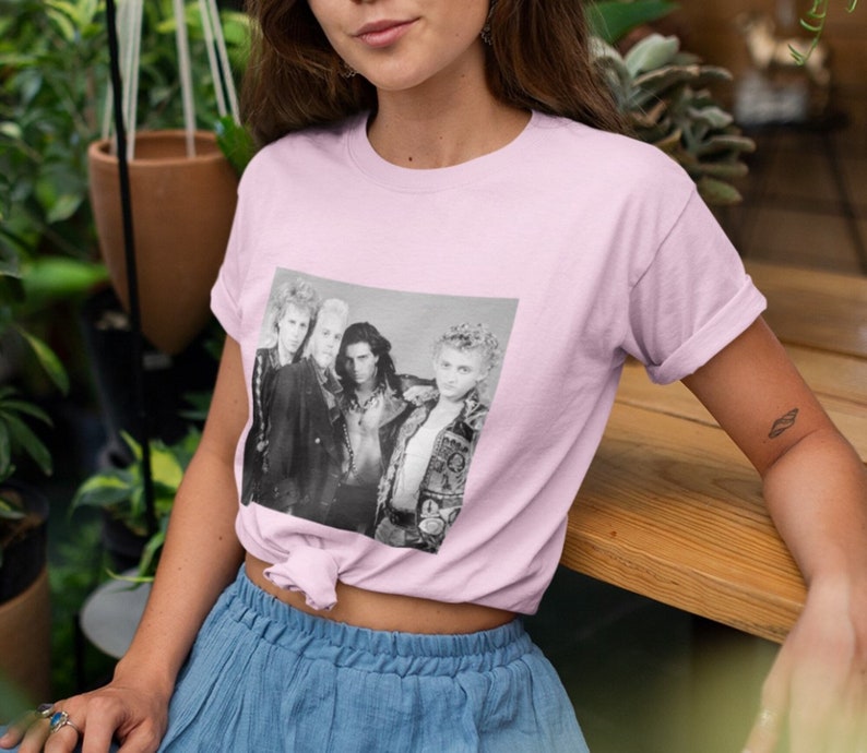 Op de afbeelding: Een roze T-shirt met een zwart-witfoto van een band op de voorkant. De bandleden dragen leren jacks en hebben lang haar. De foto is omlijst met een witte rand.