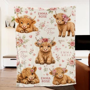 May include: A soft blanket featuring a repeating pattern of five fluffy Highland calves, pink floral arrangements, and small hearts. The name EMMA ROSE is printed in a pink serif font throughout the design. The calves are adorned with flower crowns.
