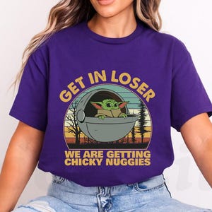 May include: Purple t-shirt featuring a graphic of a green alien character in a pod with the text "GET IN LOSER WE ARE GETTING CHICKY NUGGIES" in yellow and white. The design has a retro sunset and forest background.