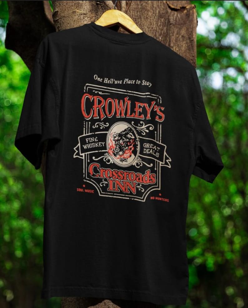 Pode incluir: Camiseta preta com um design gr&aacute;fico em estilo vintage. O design apresenta o texto "Crowley's Crossroads Inn" em vermelho e branco, com texto adicional que diz "Fine Whiskey" e "Great Deals". Uma imagem circular de um dem&ocirc;nio est&aacute; no centro.