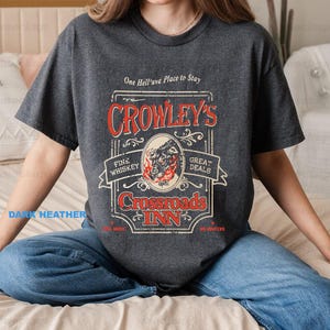 Pode incluir: T-shirt cinzento escuro com um design gr&aacute;fico vintage. O design apresenta o texto "Crowley's Crossroads Inn" em vermelho e branco, com uma ilustra&ccedil;&atilde;o de uma criatura semelhante a um lobo. A camisa tamb&eacute;m inclui o texto "Fine Whiskey" e "Great Deals".
