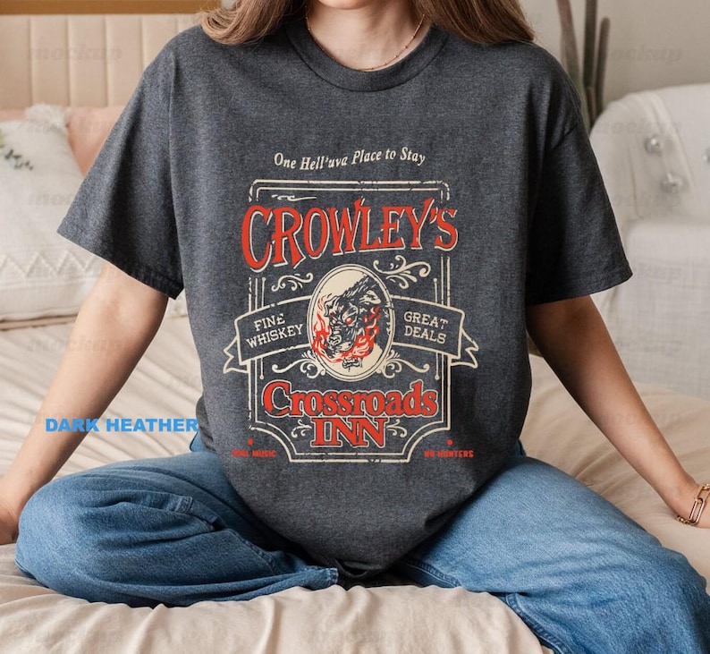 Pode incluir: T-shirt cinzento escuro com um design gr&aacute;fico vintage. O design apresenta o texto "Crowley's Crossroads Inn" em vermelho e branco, com uma ilustra&ccedil;&atilde;o de uma criatura semelhante a um lobo. A camisa tamb&eacute;m inclui o texto "Fine Whiskey" e "Great Deals".