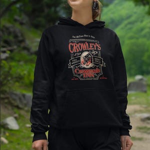 Pode incluir: Moletom preto com capuz com um gr&aacute;fico que diz "Crowley's Crossroads Inn". O design inclui as palavras "Fine Whiskey" e "Great Deals" em um estilo vintage. O moletom tem um bolso frontal e um capuz com cord&atilde;o.