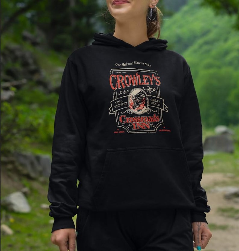 Pode incluir: Moletom preto com capuz com um gr&aacute;fico que diz "Crowley's Crossroads Inn". O design inclui as palavras "Fine Whiskey" e "Great Deals" em um estilo vintage. O moletom tem um bolso frontal e um capuz com cord&atilde;o.