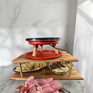 Könnte beinhalten: Ein roter elektrischer Raclette-Grill mit Holzbeinen steht auf einem zweistöckigen Serviertablett aus Holz. Das Tablett ist mit verschiedenen Lebensmitteln gefüllt, darunter geschnittener Käse, Fleisch und Gemüse.